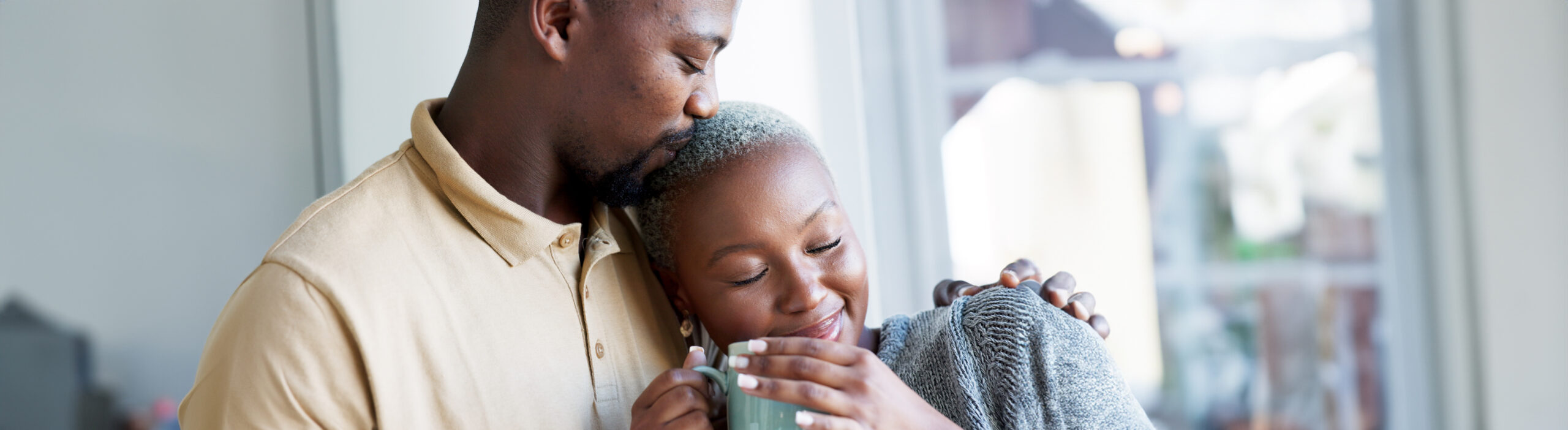 Relax, love and couple hug with coffee drink break at window to enjoy while they bond together. Kind, caring and happy boyfriend giving black woman beverage with romantic embrace in home.